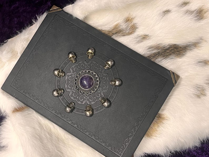 May include: A black leather-bound book with decorative corner protectors. The cover features a central silver medallion with a purple stone, surrounded by a circle of small silver skulls. The book rests on a white and brown fur-like surface.