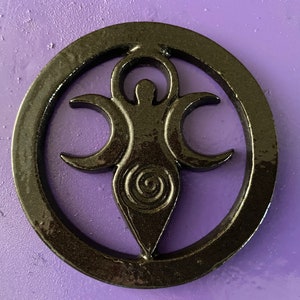 May include: Black metal wall hanging with a triple moon goddess symbol. The symbol is a stylized female figure with three crescent moons around her head and a spiral design in the center.