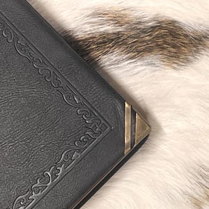 May include: A dark gray book with ornate corner detailing and a brass corner protector. The book's cover has a textured appearance. The background is a mix of white and brown fur.