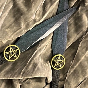 May include: Two black throwing knives with gold pentagram accents on a brown velvet background.
