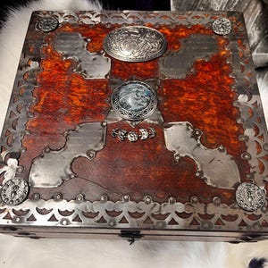 May include: A wooden box with a reddish-brown finish, featuring silver-toned metal accents. The box is decorated with metal plates, ornate corner details, and a central oval embellishment. The lid is further embellished with circular and oval decorative elements.