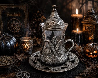 May include: A clear glass tea cup with a handle, featuring an intricate silver-toned metal holder and a matching lid. The cup rests on a circular silver-toned metal saucer with detailed floral engravings. The metal components have a dark, aged finish.