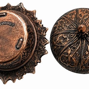 Puede incluir: Un recipiente de metal color cobre con tapa, que presenta intrincados patrones de hojas en relieve y un remate puntiagudo. La parte inferior de la base muestra el texto Made in TURKEY e incluye tres peque&ntilde;as patas para mayor estabilidad.