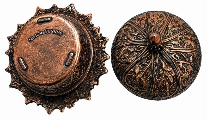 Puede incluir: Un recipiente de metal color cobre con tapa, que presenta intrincados patrones de hojas en relieve y un remate puntiagudo. La parte inferior de la base muestra el texto Made in TURKEY e incluye tres peque&ntilde;as patas para mayor estabilidad.
