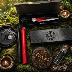 May include: A ritual set featuring a dagger with a black handle and red blade in a box, a black and red candle, a silver pendant necklace in a dish, a black box with a circular sigil, a jar of black salt, and two small lit candles.