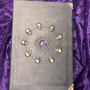 May include: A dark gray book with ornate gold corner protectors and a decorative border. The cover features a central design with a purple stone surrounded by small skull accents. The book is set against a purple velvet background.