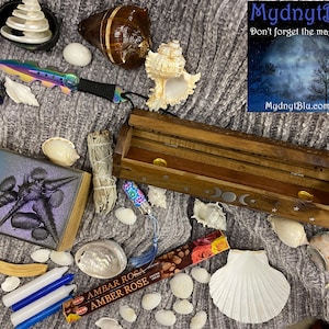 May include: A wooden incense burner with a moon and star design, surrounded by seashells, white candles, a sage smudge stick, a book with a blue and gold cover, a small knife, and a box of incense sticks labeled "AMBAR ROSA AMBER ROSE".