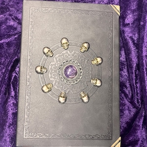 May include: A dark gray book with gold corner protectors and a decorative border. The cover features a central design with a purple stone surrounded by small skull accents.