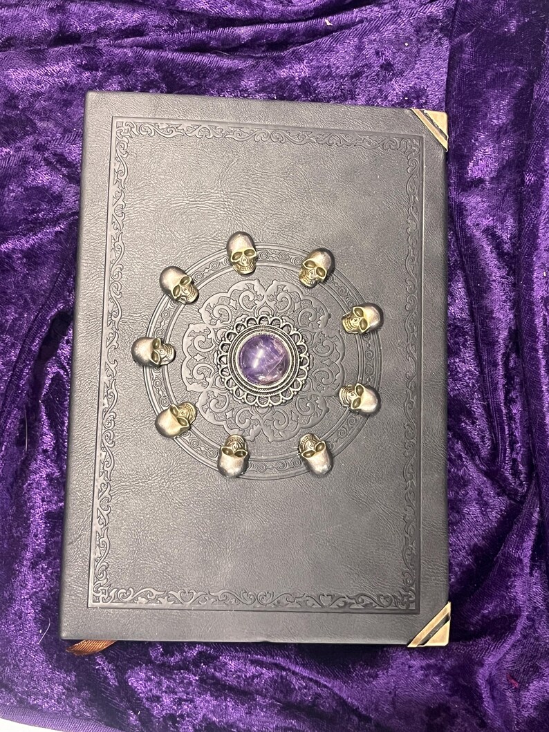 May include: A dark gray book with gold corner protectors and a decorative border. The cover features a central design with a purple stone surrounded by small skull accents.