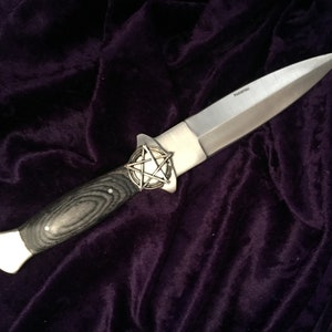 May include: A silver knife with a black wood handle and a pentagram design on the pommel. The knife is lying on a dark purple velvet surface.