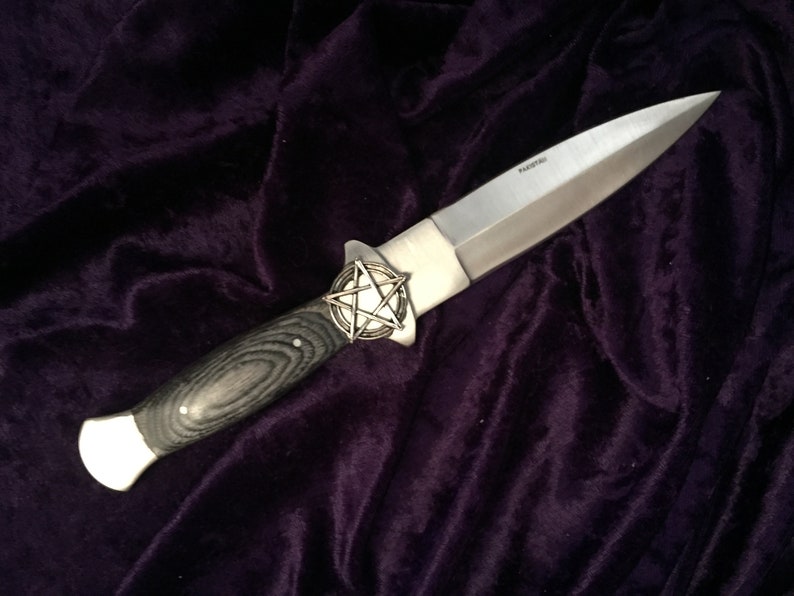 May include: A silver knife with a black wood handle and a pentagram design on the pommel. The knife is lying on a dark purple velvet surface.