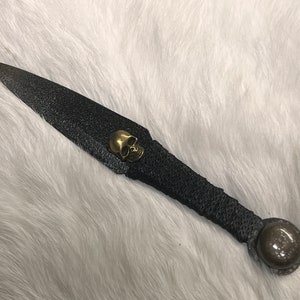 May include: A black throwing knife with a skull-shaped pommel and a black leather wrap. The knife has a textured blade and a round, black metal cap on the end of the handle.
