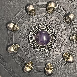 May include: A decorative, dark gray journal with a detailed embossed pattern. The journal is adorned with small silver skull embellishments arranged in a circle around a central silver pendant with a purple stone.
