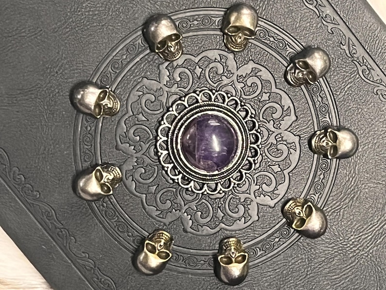 May include: A decorative, dark gray journal with a detailed embossed pattern. The journal is adorned with small silver skull embellishments arranged in a circle around a central silver pendant with a purple stone.