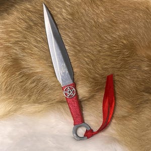 May include: A silver-colored dagger with a pointed blade and a red, woven handle. A silver pentagram is on the handle. A red ribbon is attached to the handle. The dagger is lying on a brown and white fur surface.