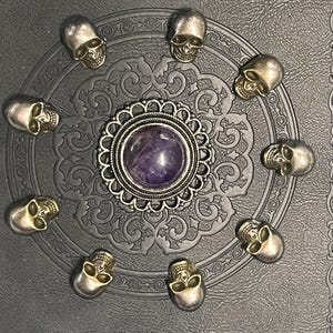 May include: A dark gray decorative piece featuring a central purple stone surrounded by a silver filigree design. Ten small silver skull embellishments encircle the center, creating a gothic or Halloween theme.