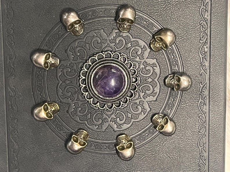May include: A dark gray decorative piece featuring a central purple stone surrounded by a silver filigree design. Ten small silver skull embellishments encircle the center, creating a gothic or Halloween theme.