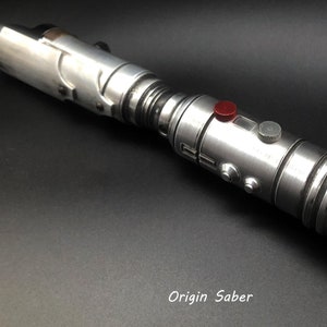 Puede incluir: Un sable de luz plateado con detalles negros y un bot&oacute;n rojo. El sable de luz tiene un anillo en el extremo y el texto "Origin Saber" est&aacute; impreso debajo del sable de luz.