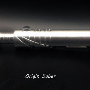 May include: A silver lightsaber with a metallic finish. The hilt has buttons and a textured grip. The blade emitter is angled, and the design suggests a futuristic weapon. The text "Origin Saber" is printed at the bottom.