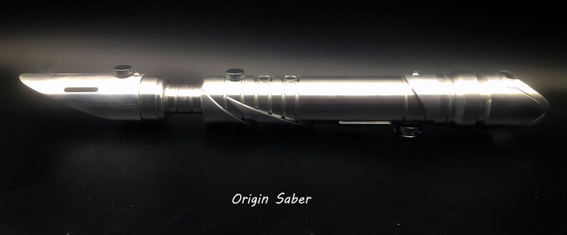 May include: A silver lightsaber with a metallic finish. The hilt has buttons and a textured grip. The blade emitter is angled, and the design suggests a futuristic weapon. The text "Origin Saber" is printed at the bottom.