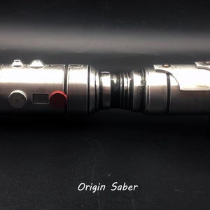 Puede incluir: Un sable de luz plateado con un bot&oacute;n rojo en el lateral. El sable de luz est&aacute; sobre una superficie negra y el texto "Origin Saber" es visible debajo del sable de luz.