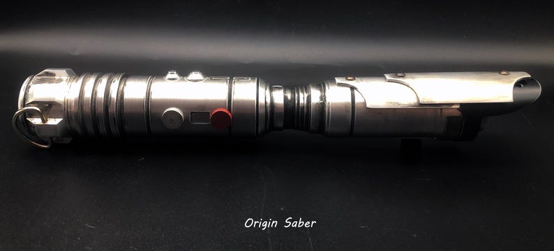 Puede incluir: Un sable de luz plateado con un bot&oacute;n rojo en el lateral. El sable de luz est&aacute; sobre una superficie negra y el texto "Origin Saber" es visible debajo del sable de luz.