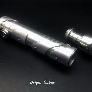 May include: Three disassembled silver lightsaber components on a black surface. The parts include a cylindrical body, a hilt, and a blade emitter. The text "Origin Saber" is visible.