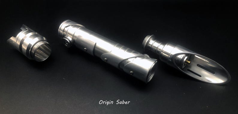 May include: Three disassembled silver lightsaber components on a black surface. The parts include a cylindrical body, a hilt, and a blade emitter. The text "Origin Saber" is visible.