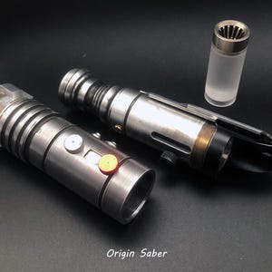 Puede incluir: Dos empu&ntilde;aduras de sable l&aacute;ser plateadas con detalles negros y un peque&ntilde;o tubo transparente con tapa plateada. Las empu&ntilde;aduras tienen una superficie texturizada y un bot&oacute;n en el lateral. El texto "Origin Saber" est&aacute; impreso en la parte inferior de la imagen.