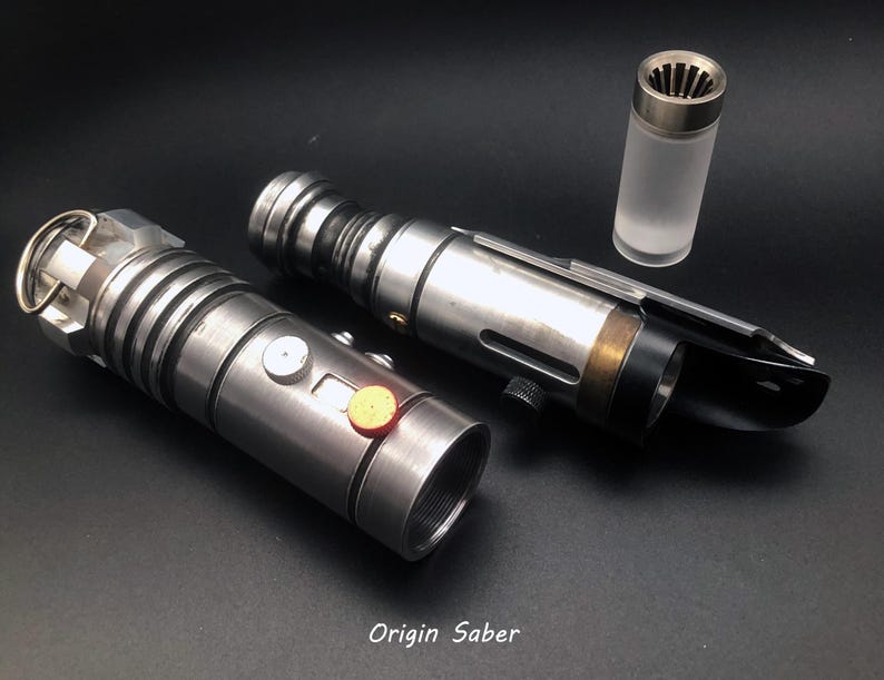 Puede incluir: Dos empu&ntilde;aduras de sable l&aacute;ser plateadas con detalles negros y un peque&ntilde;o tubo transparente con tapa plateada. Las empu&ntilde;aduras tienen una superficie texturizada y un bot&oacute;n en el lateral. El texto "Origin Saber" est&aacute; impreso en la parte inferior de la imagen.