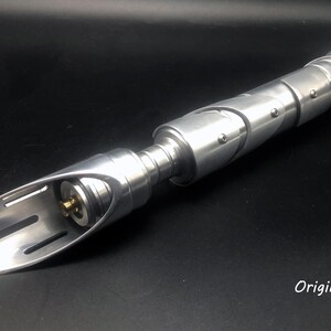 May include: A silver lightsaber with a curved blade and a segmented handle. The handle has visible screws and a circular button. The text "Origin Saber" is visible in the lower right corner.