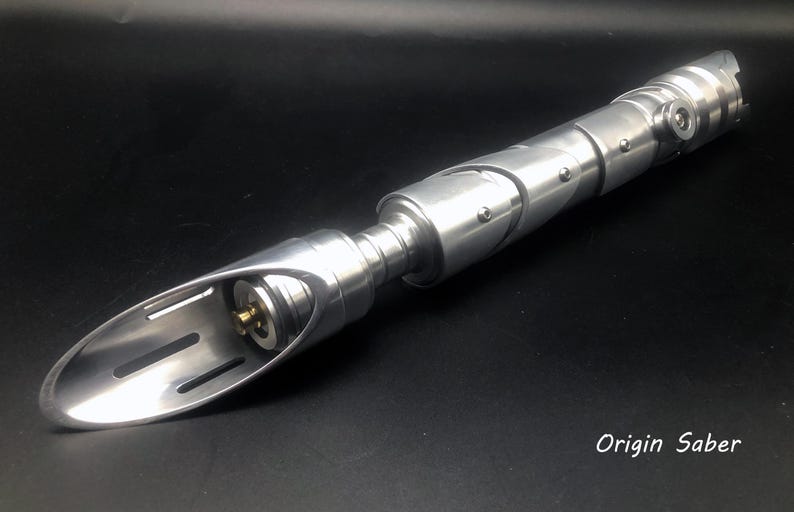 May include: A silver lightsaber with a curved blade and a segmented handle. The handle has visible screws and a circular button. The text "Origin Saber" is visible in the lower right corner.