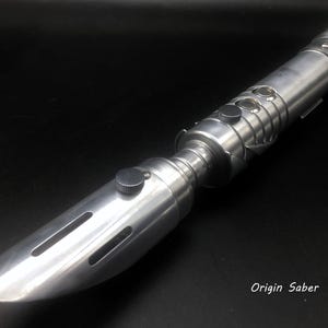 May include: A silver lightsaber with a rounded tip and multiple buttons and ridges. The lightsaber has a cylindrical body with a textured grip against a black background. The text "Origin Saber" is visible.