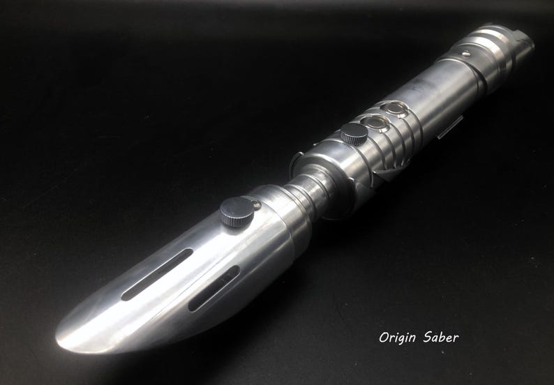May include: A silver lightsaber with a rounded tip and multiple buttons and ridges. The lightsaber has a cylindrical body with a textured grip against a black background. The text "Origin Saber" is visible.