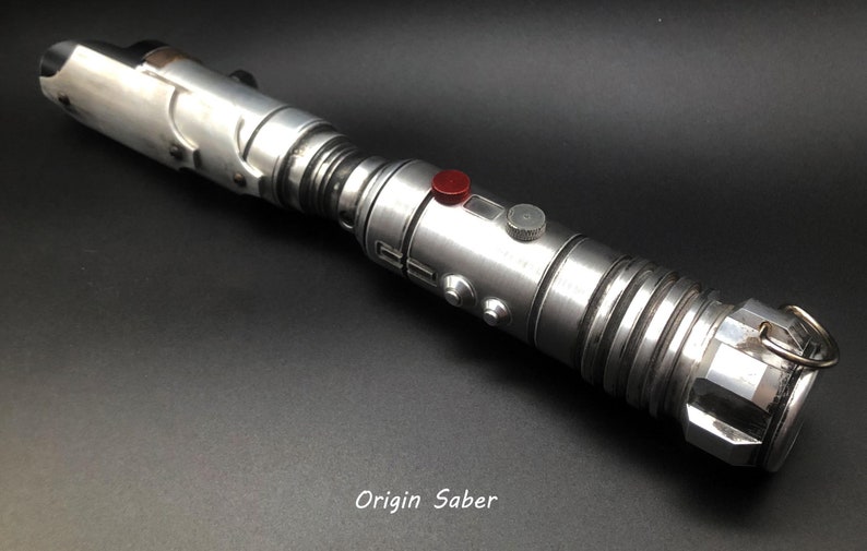 Puede incluir: Un sable de luz plateado con detalles negros y un bot&oacute;n rojo. El sable de luz tiene un anillo en el extremo y el texto "Origin Saber" est&aacute; impreso debajo del sable de luz.