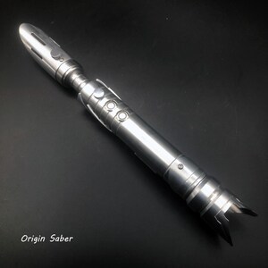 May include: A metallic silver lightsaber with a detailed design. The lightsaber features a cylindrical body, buttons, and a blade emitter. The text "Origin Saber" is visible at the bottom of the image.