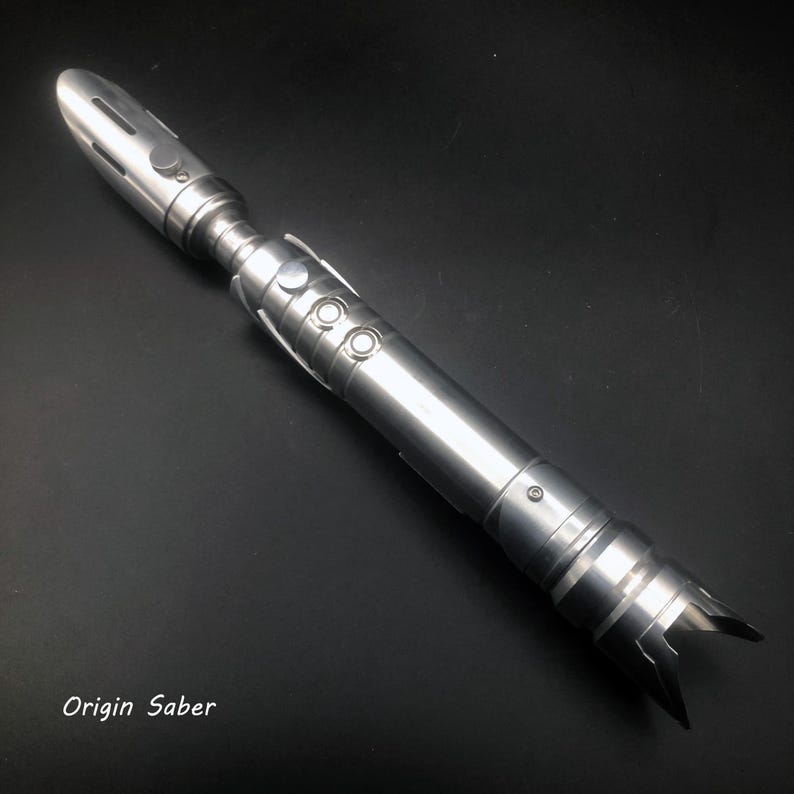 May include: A metallic silver lightsaber with a detailed design. The lightsaber features a cylindrical body, buttons, and a blade emitter. The text "Origin Saber" is visible at the bottom of the image.