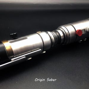Puede incluir: Un sable de luz de utiler&iacute;a plateado con detalles en negro y dorado. El sable de luz tiene un bot&oacute;n rojo y un mango negro. El texto "Origin Saber" est&aacute; impreso en la superficie negra debajo del sable de luz.