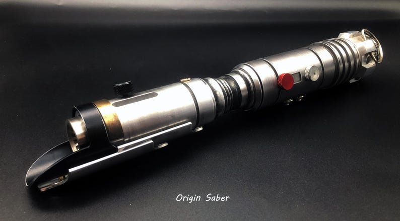 Puede incluir: Un sable de luz de utiler&iacute;a plateado con detalles en negro y dorado. El sable de luz tiene un bot&oacute;n rojo y un mango negro. El texto "Origin Saber" est&aacute; impreso en la superficie negra debajo del sable de luz.