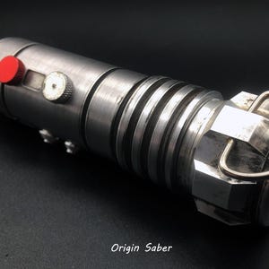 Puede incluir: Un sable de luz plateado con detalles negros y un bot&oacute;n rojo. El sable de luz tiene un anillo de metal en el extremo.