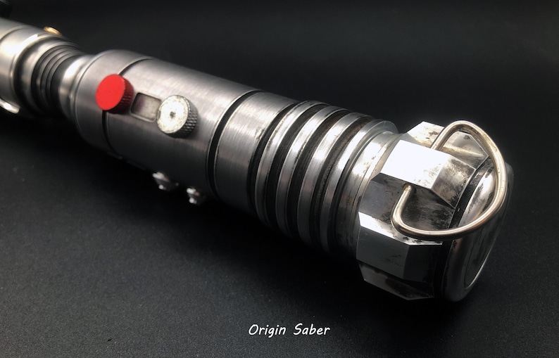 Puede incluir: Un sable de luz plateado con detalles negros y un bot&oacute;n rojo. El sable de luz tiene un anillo de metal en el extremo.