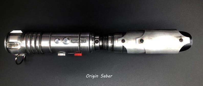 Puede incluir: Un sable de luz plateado con un mango negro y un bot&oacute;n rojo. El sable de luz tiene una carcasa de metal plateado con un agarre negro y un bot&oacute;n rojo en el lateral. El sable de luz es un juguete popular para los fan&aacute;ticos de la franquicia Star Wars.