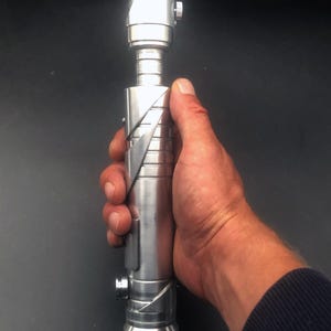 May include: A silver lightsaber with a blade-like top and a detailed handle. The lightsaber is held in a hand against a dark background. The text "Origin Saber" is in the bottom left corner.