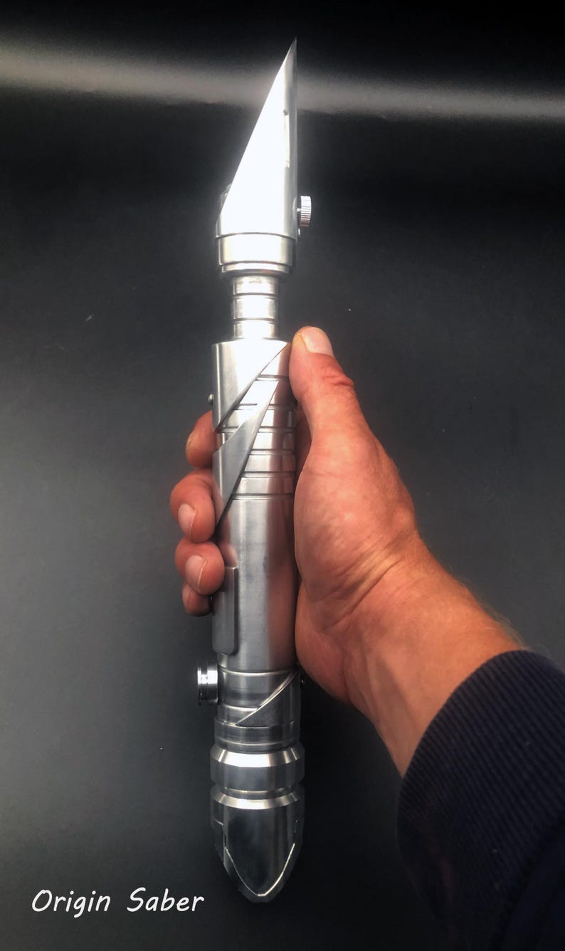 May include: A silver lightsaber with a blade-like top and a detailed handle. The lightsaber is held in a hand against a dark background. The text "Origin Saber" is in the bottom left corner.
