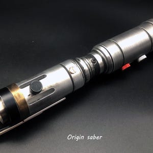 Puede incluir: Un sable de luz plateado con detalles en negro y dorado. El sable de luz tiene un bot&oacute;n rojo y un interruptor negro. El sable de luz est&aacute; sobre una superficie negra. Origin saber.