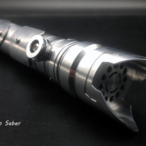 May include: A silver lightsaber with a detailed, metallic design. The hilt features segmented sections, rivets, and a flared emitter with circular cutouts. The text "Origin Saber" is visible.