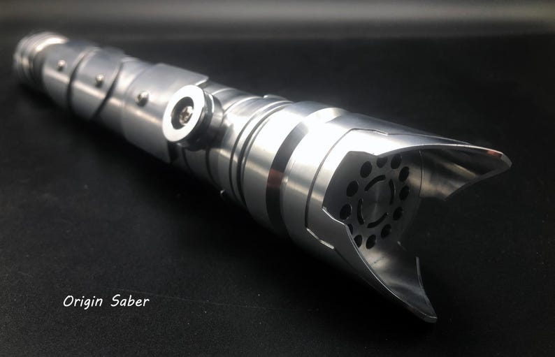 May include: A silver lightsaber with a detailed, metallic design. The hilt features segmented sections, rivets, and a flared emitter with circular cutouts. The text "Origin Saber" is visible.
