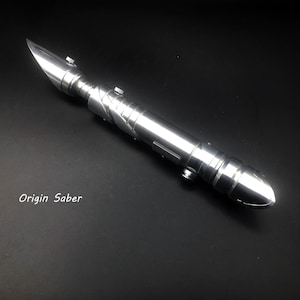 May include: A silver lightsaber with a pointed blade and a detailed handle. The lightsaber has a metallic finish and is lying on a black surface. The text "Origin Saber" is visible.