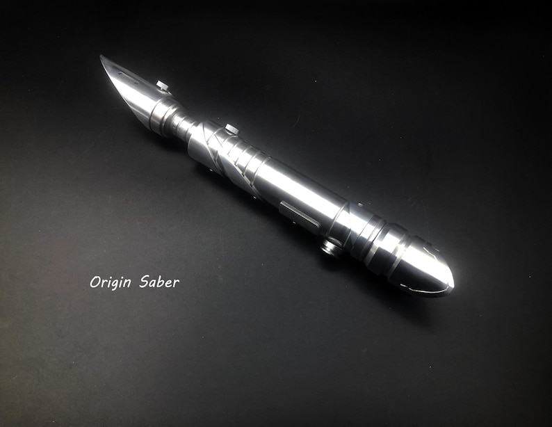 May include: A silver lightsaber with a pointed blade and a detailed handle. The lightsaber has a metallic finish and is lying on a black surface. The text "Origin Saber" is visible.