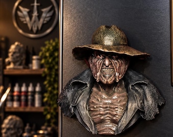 May include: A three-dimensional refrigerator magnet depicting a character with a scarred, textured face, wearing a wide-brimmed hat and a dark, tattered jacket. The figure has a menacing expression with visible teeth and a weathered, bronze-toned finish.